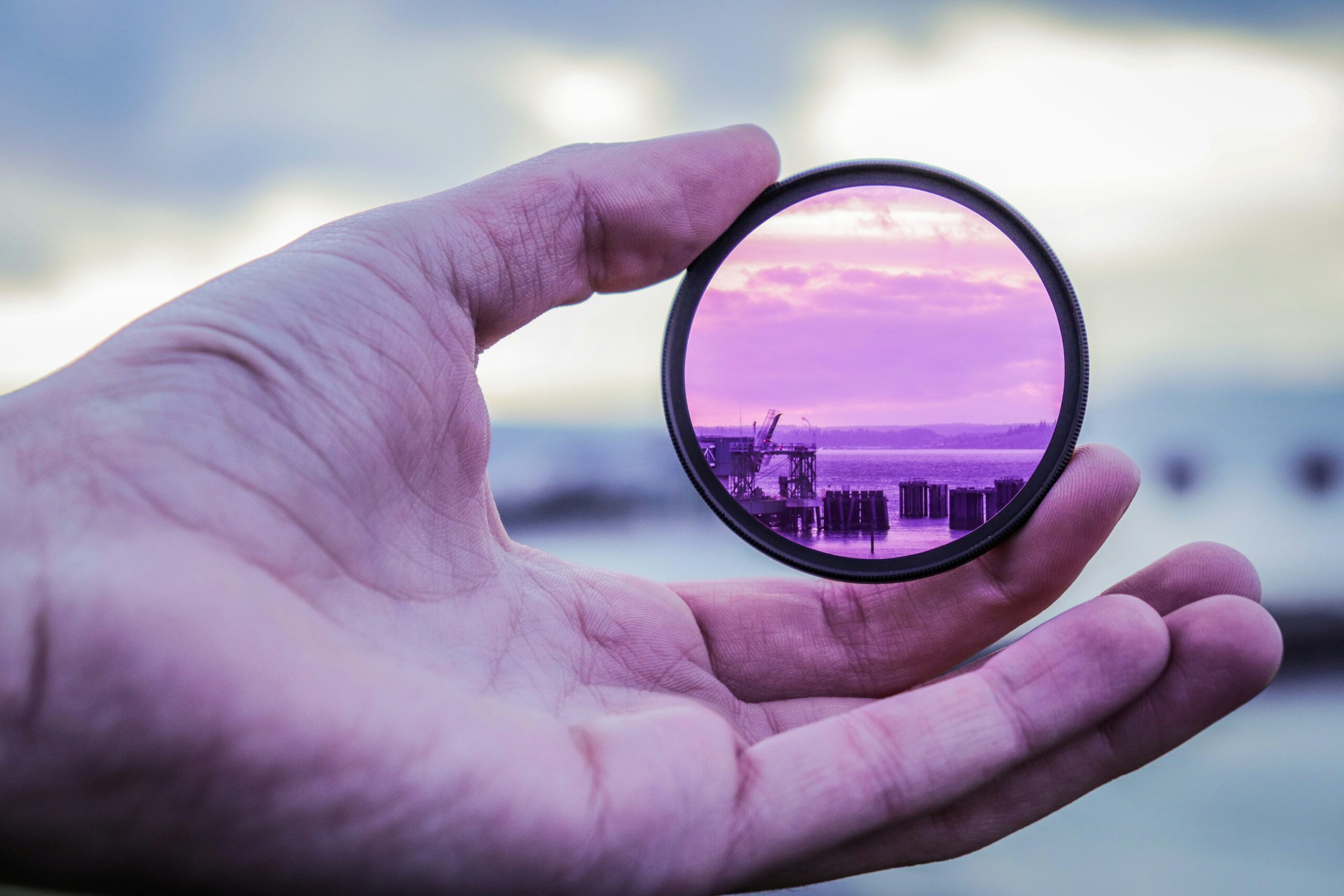 A hand holding a circular filter, with a purple hue, framing a view of a waterfront scene.