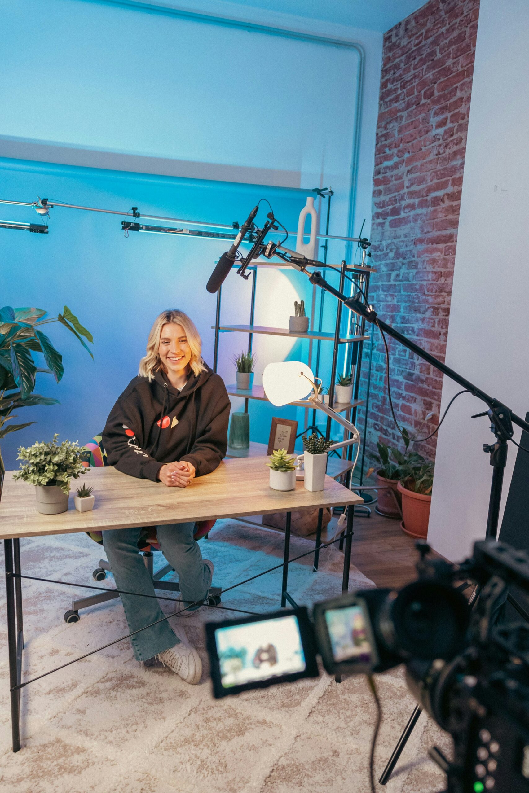 A smiling individual wearing a black hoodie sits at a desk surrounded by potted plants and filming equipment in a well-lit studio.
